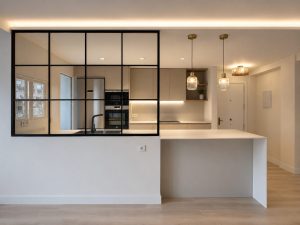Salón con cerramiento de acero y vidrio hacia la cocina – reforma integral de vivienda en Torrelodones